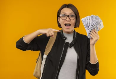 impressed-young-student-girl-wearing-glasses-back-bag-holding-pointing-money-isolated-orange